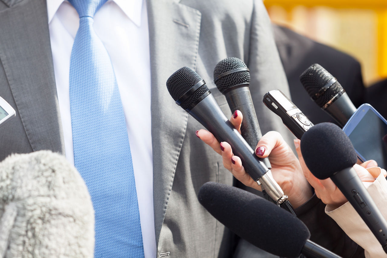 person in suit and blue tie taking questions from multiple microphones